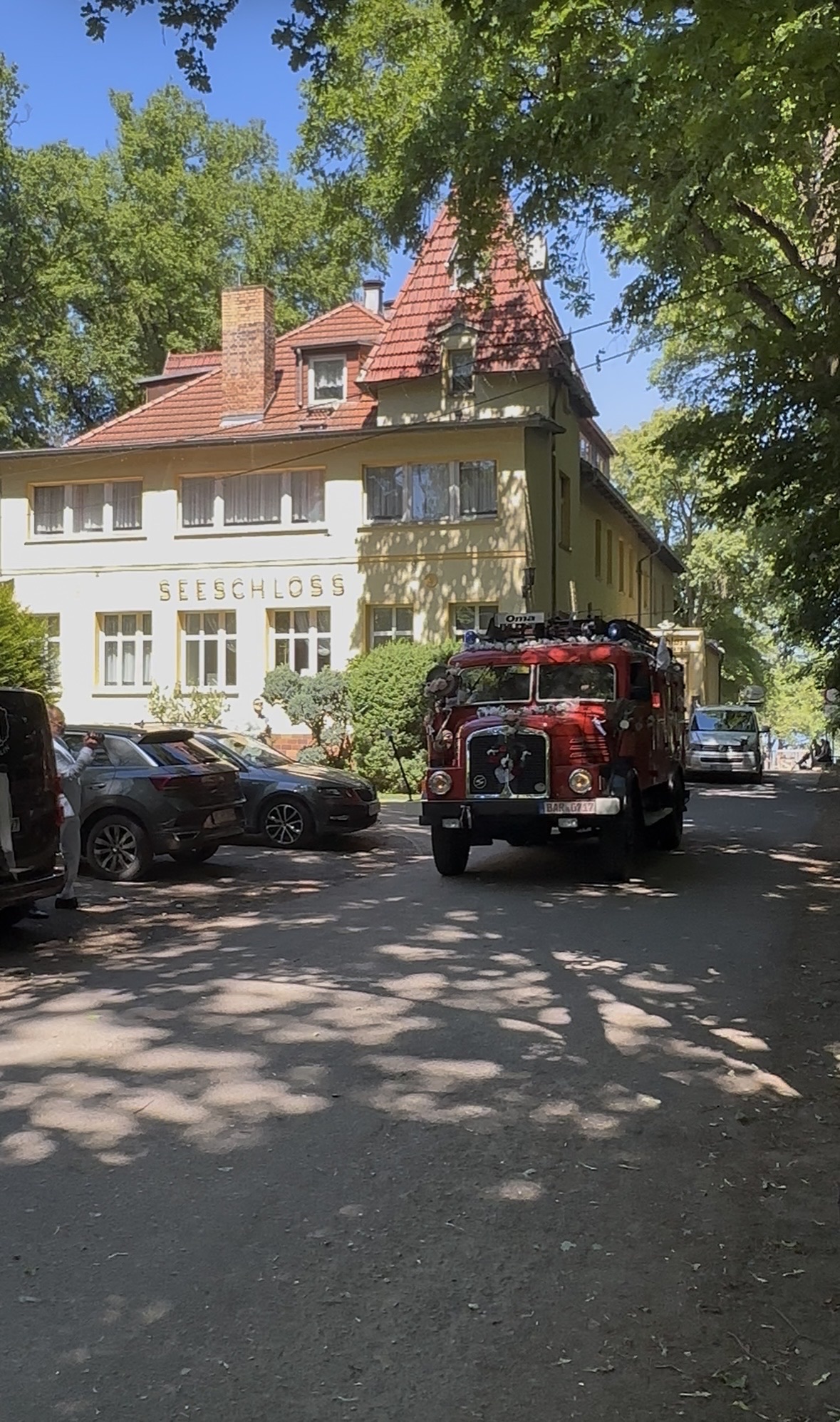 Historisches rotes Feuerwehrfahrzeug als Hochzeitsauto vor dem Hotel Seeschloss am Bötzsee in Petershagen-Eggersdorf. Das dekorierte Fahrzeug fährt zur Hochzeit vor, umgeben von alten Bäumen und der romantischen Kulisse der Hochzeitslocation am See.