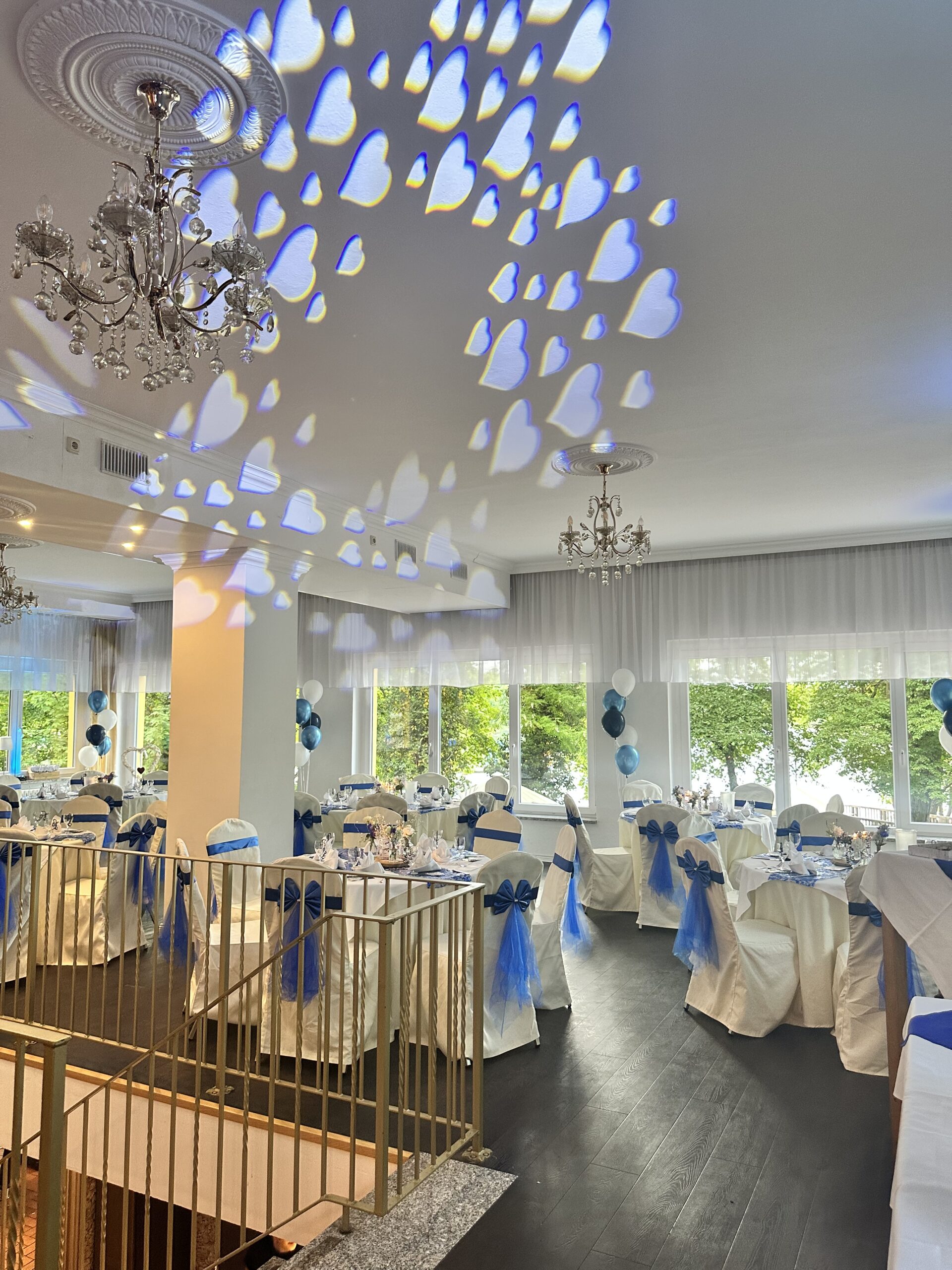 Festlich dekorierter Panoramasaal im Hotel Seeschloss am Bötzsee mit weiß-blauer Hochzeitsdekoration, eingedeckten Tischen, Blick auf den See und romantischen Herz-Lichtreflexen an der Decke.