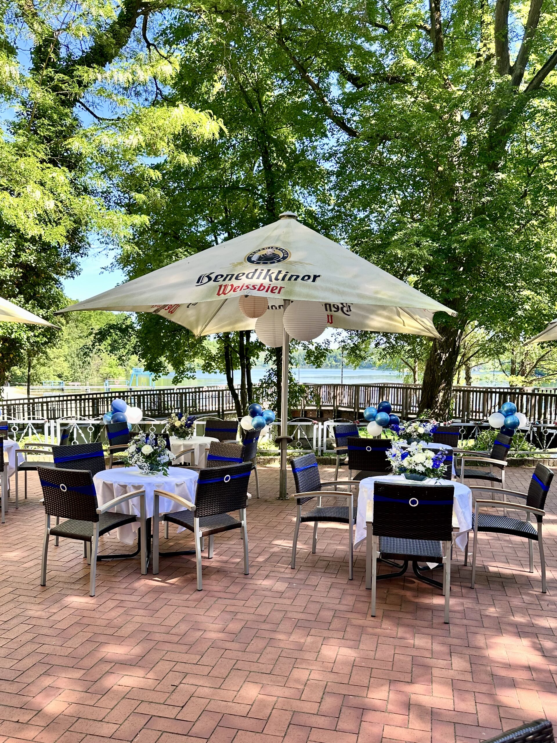 Festlich gedeckte Seeterrasse im Hotel Seeschloss am Bötzsee mit weiß-blauer Hochzeitsdekoration, Kaffeetafeln, Sonnenschirmen und direktem Blick auf den See.