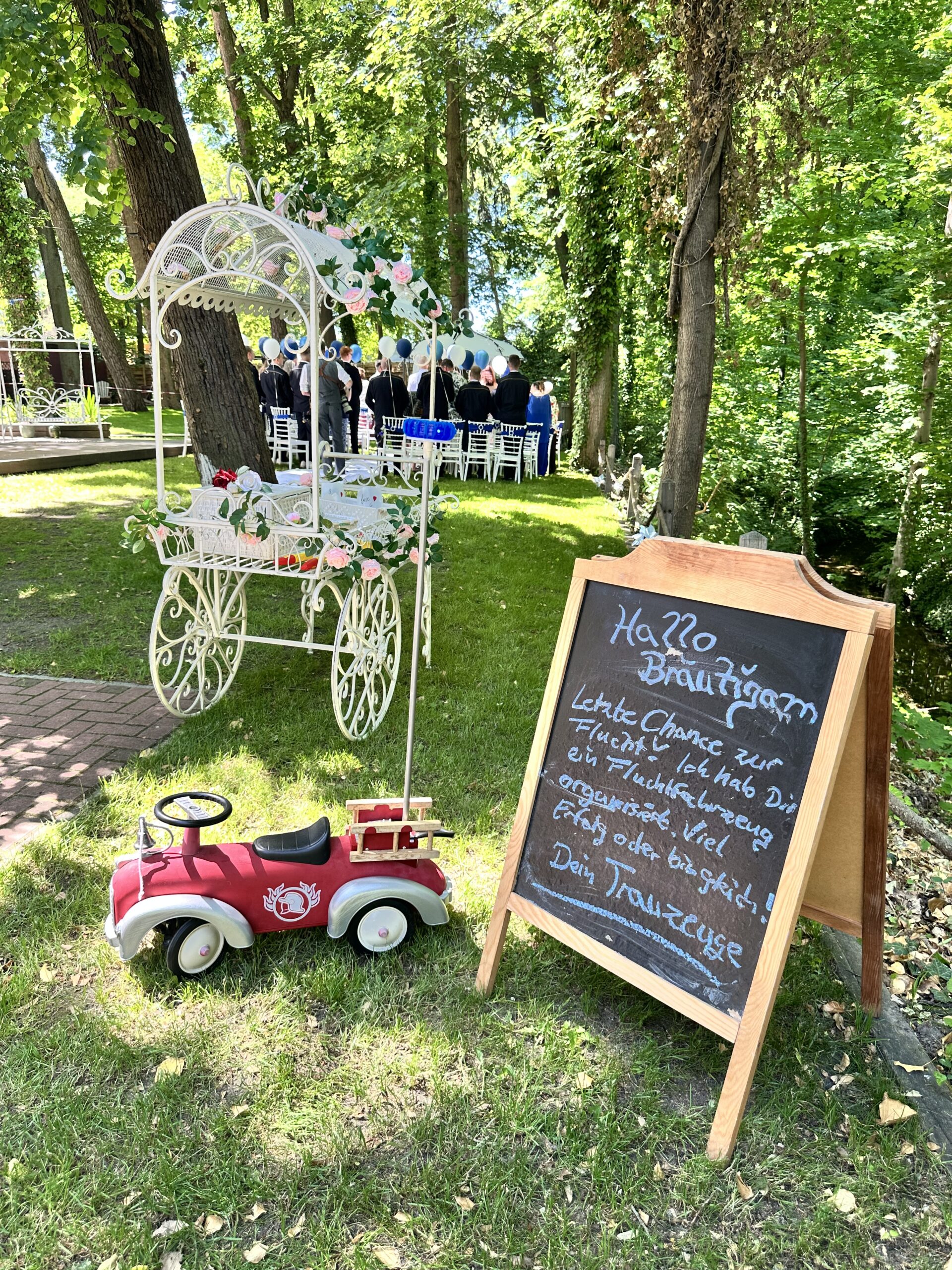 Liebevoll dekorierter Trauungsbereich im Garten des Hotel Seeschloss am Bötzsee mit Willkommensschild für den Bräutigam, dekorativem Blumenwagen und Hochzeitsgästen während einer standesamtlichen Trauung unter alten Bäumen.